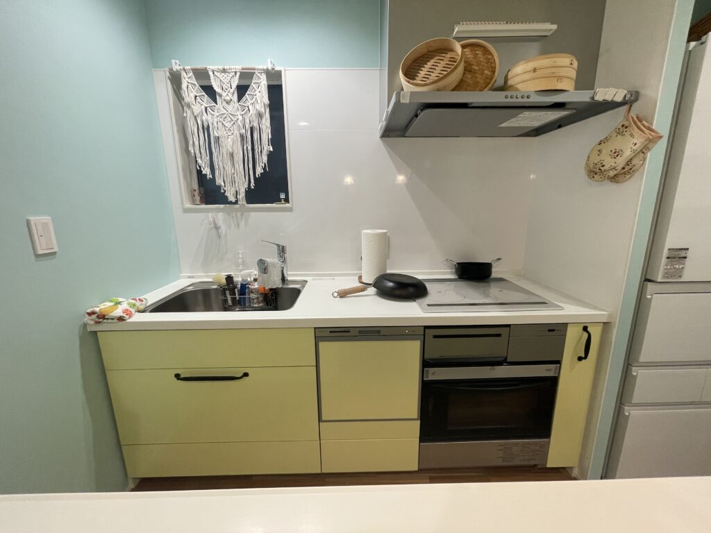 白い天板と黄色のキッチン。左にシンク、右にIHコンロ。ビルドイン食洗機とビルドインオーブンレンジがある。シンクの前の型ガラスにはマクラメが掛かっている。